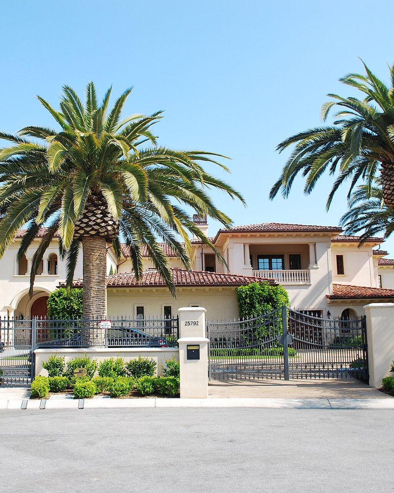 A large tiled roof house behind an iron gate and two tall palm trees, the street number set on the gatepost